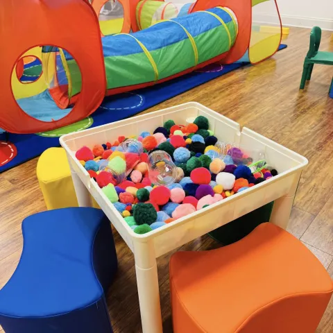 Play children's tunnels in the background. A sensory bin table with colorful pom poms is in the front.